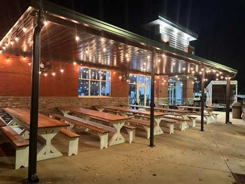 Outdoor dining area with wooden tables, string lights, and a brick building, illuminated at night.