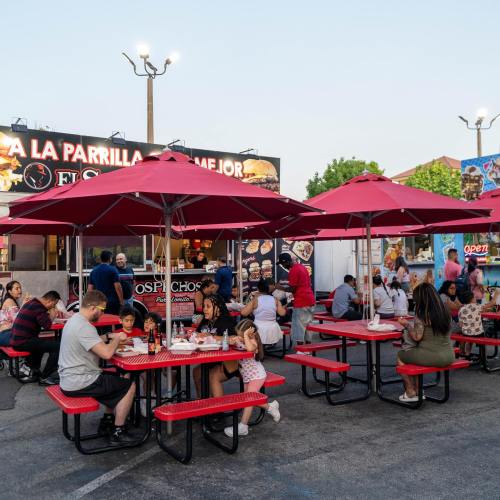 A lively outdoor dining area with red umbrellas, families enjoying food at picnic tables, and food trucks in the background.