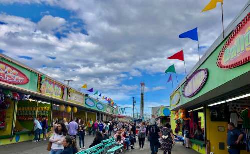 A bustling fairground with colorful stalls, people walking, and flags waving under a cloudy sky.