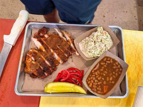A tray with sliced grilled chicken in sauce, coleslaw, baked beans, pickles, and red onions on a paper liner.