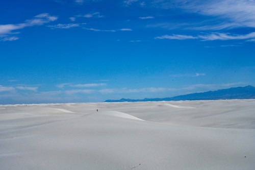 A vast desert landscape with rolling white sand dunes under a clear blue sky and distant mountains.
