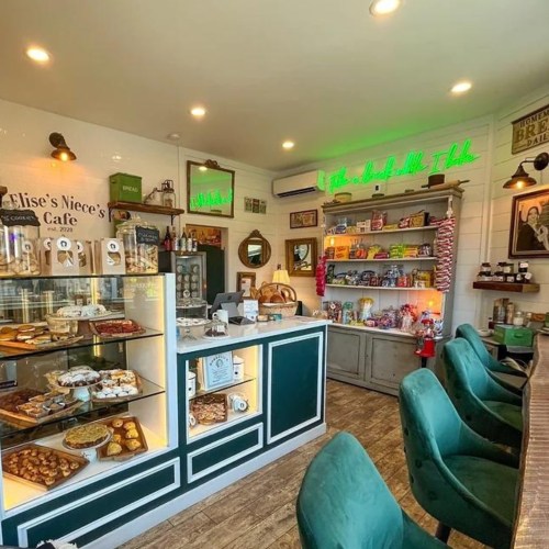 A cozy café interior featuring a display of baked goods, shelves of snacks, and green neon signage.
