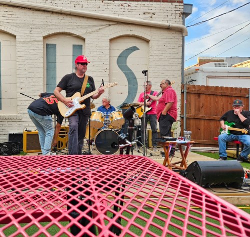 A band performs outdoors with a red table in the foreground, featuring a guitarist, drummer, and other musicians.