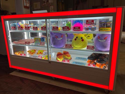 A brightly lit prize display case featuring plush toys, snacks, and game tickets, with vibrant red accents.