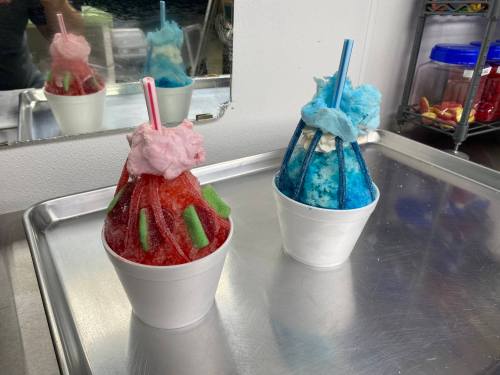 Two colorful snow cones in white cups, one red with green toppings and cotton candy, the other blue with a cotton candy top.