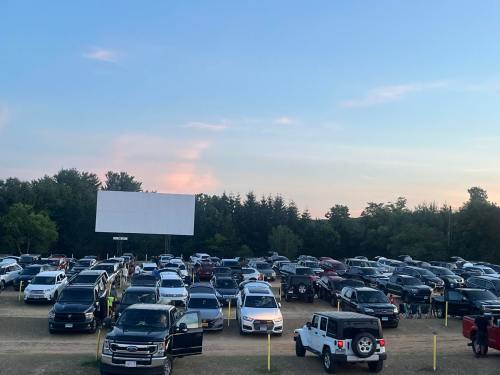 A crowded outdoor parking lot filled with cars, with a large movie screen in the background under a clear sky.