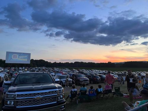 A crowded drive-in theater at sunset, with cars parked and a large screen displaying a movie.