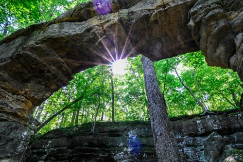 Sunlight beams through a natural stone arch, surrounded by lush green trees and rocky terrain.