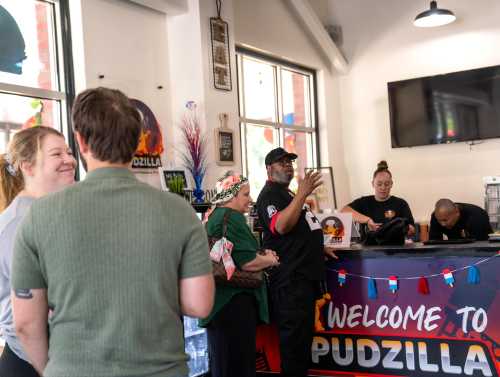 A busy restaurant scene with customers interacting and staff at the counter, featuring a colorful "Welcome to Pudzilla" sign.