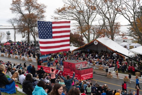 A large crowd gathers at an outdoor event with a giant American flag and a stage under a tent in the background.