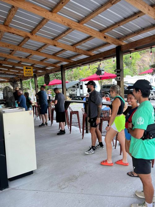 A group of people waiting in line at an outdoor food stand with picnic tables and RVs in the background.