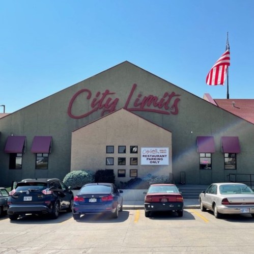 Building with "City Limits" sign, American flag, and parking area with several cars in front. Clear blue sky above.