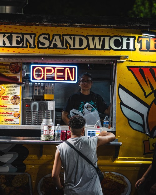 A food truck with a glowing "OPEN" sign, serving chicken sandwiches, as a customer places an order.