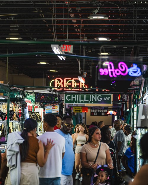 A bustling market scene with people browsing shops, colorful signs, and a vibrant atmosphere.