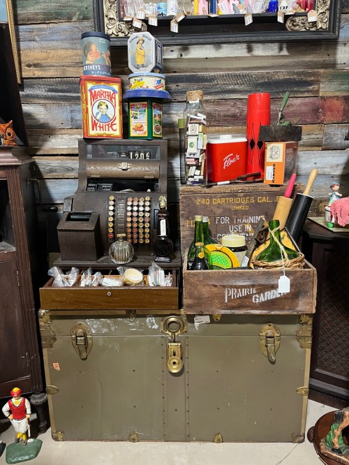 A vintage display featuring an old cash register, wooden crates, bottles, and various nostalgic items on a rustic backdrop.