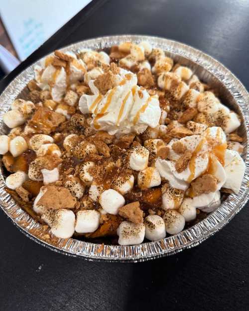 A dessert pie topped with whipped cream, toasted marshmallows, crushed graham crackers, and caramel drizzle.