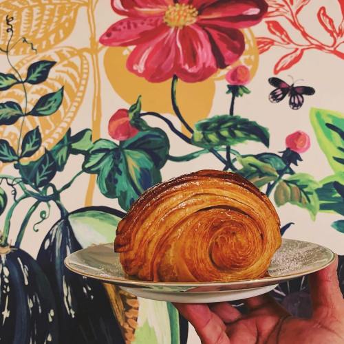 A hand holds a golden, flaky croissant on a plate in front of a vibrant floral mural.