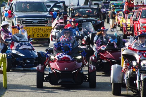 A parade of decorated trikes and vehicles, featuring riders in patriotic attire with flags and red, white, and blue decorations.