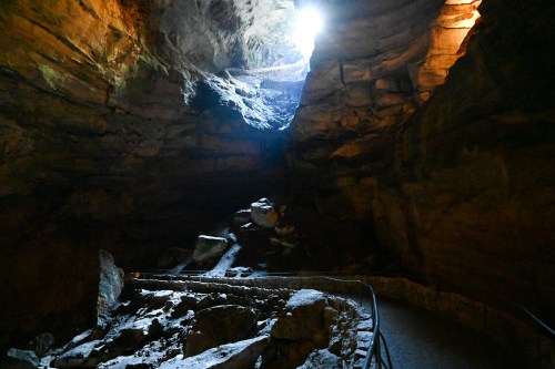A dimly lit cave with rocky formations and a bright opening at the top, revealing sunlight streaming in.