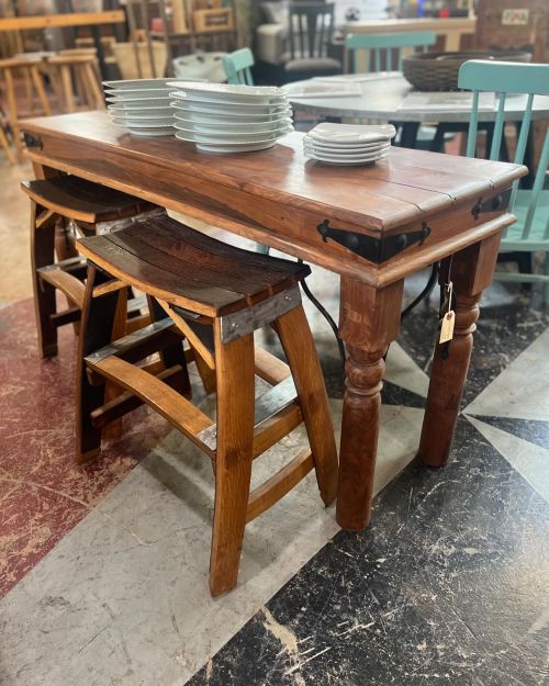 A wooden table with stacked plates on top, surrounded by various chairs and stools in a rustic setting.
