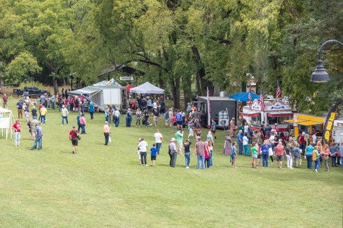 A crowd gathers at an outdoor event with food stalls and tents set up among trees on a grassy area.