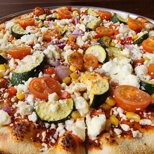 A close-up of a pizza topped with tomatoes, zucchini, corn, red onion, and crumbled cheese on a golden crust.