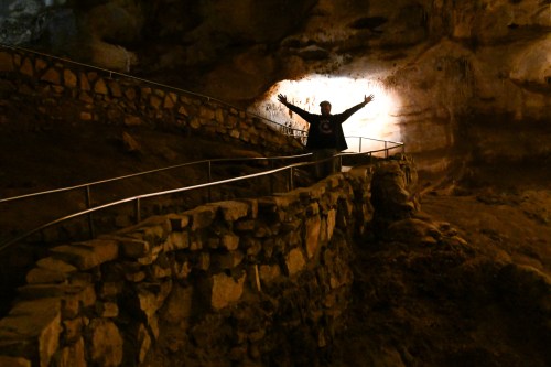 A person stands with arms outstretched in a cave, illuminated by light, with stone pathways and walls surrounding them.