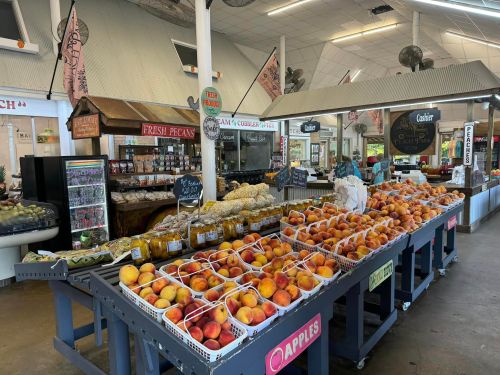 A vibrant market stall filled with fresh peaches, pecans, and various local products in a spacious indoor setting.