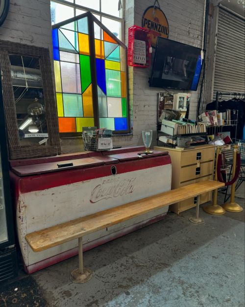 A vintage Coca-Cola cooler with a wooden bench in front, beside a colorful stained glass window and shelves of books.