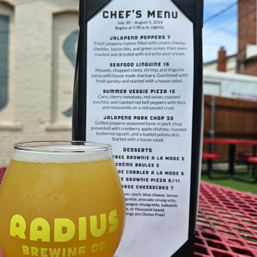 A close-up of a beer glass in front of a menu featuring various dishes at Radius Brewing Co. on a red table.