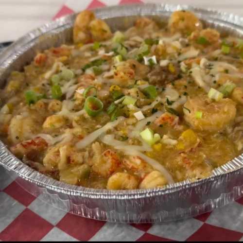 A foil container filled with shrimp and seafood gumbo, topped with green onions and served on a checkered paper background.