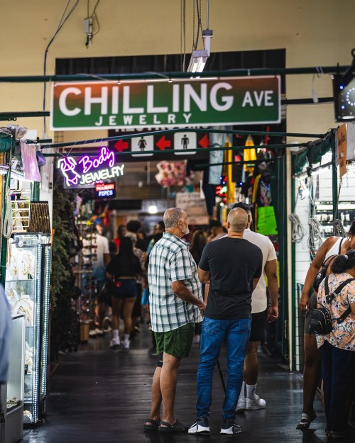 A bustling market corridor with shoppers, featuring a sign for "Chilling Ave Jewelry" and colorful displays.