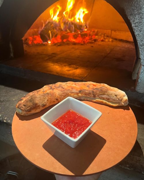 A freshly baked calzone on a wooden plate, accompanied by a small bowl of red sauce, with a wood-fired oven in the background.