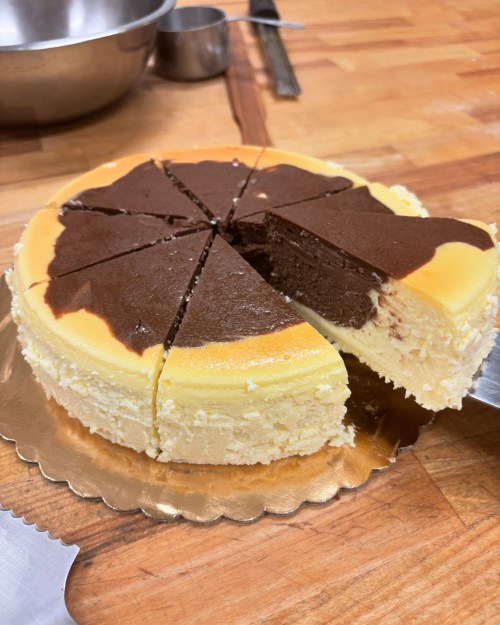 A sliced cheesecake with a chocolate layer on top, resting on a wooden surface with baking tools in the background.