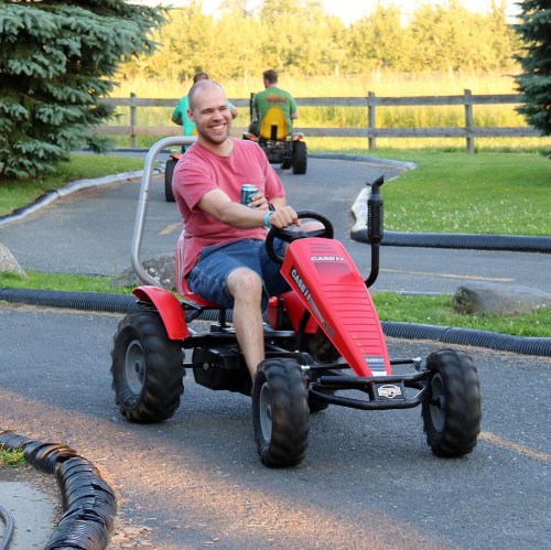 A man joyfully rides a red pedal-powered go-kart on a track, with trees and a grassy field in the background.