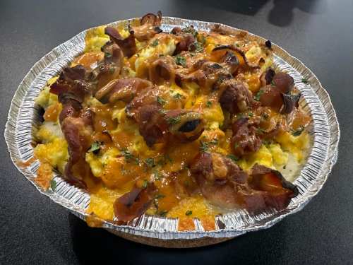 A foil tray filled with scrambled eggs, melted cheese, and crispy bacon, garnished with herbs.