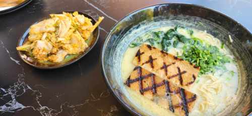 A bowl of creamy ramen topped with grilled tofu and spinach, alongside a small dish of kimchi.