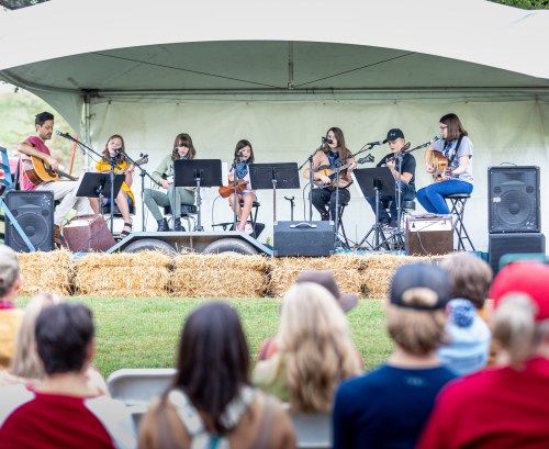 A group of young musicians performs on stage under a tent, playing various instruments for an audience.