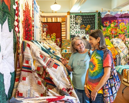 Two women admire colorful quilts displayed in a vibrant quilt show, surrounded by various intricate designs.