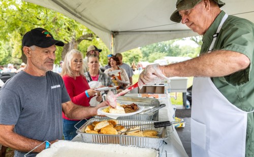 A man receives a plate of food from a server at an outdoor event, with others waiting in line under a tent.