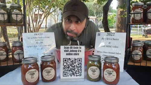 A man at a market stall displays jars of salsa and a QR code for visiting his online store.