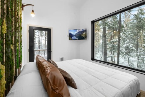 Cozy bedroom with a large window showing a snowy forest, featuring a bed with brown pillows and a TV on the wall.