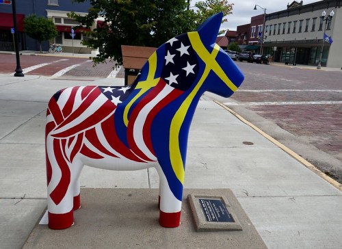A colorful statue of a horse decorated with American and Swedish flag patterns, located on a city sidewalk.