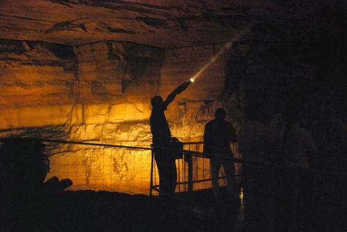 A person points a flashlight in a dimly lit cave, illuminating the rocky walls while others observe nearby.