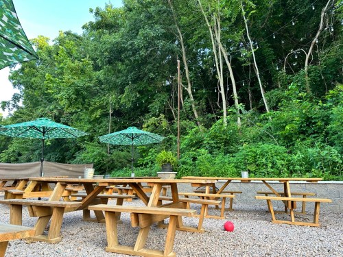 Outdoor seating area with wooden picnic tables, green umbrellas, and lush greenery in the background.
