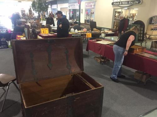 A vintage market scene with people browsing antiques, including an open wooden chest and various displayed items.