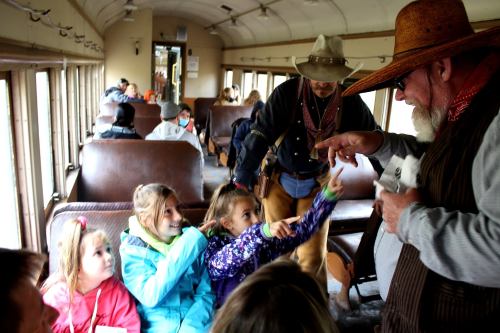 A group of children interact with a costumed performer on a vintage train, while adults observe in the background.