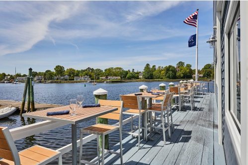 Outdoor dining area by a calm waterfront, featuring tables and chairs with flags waving in the breeze.