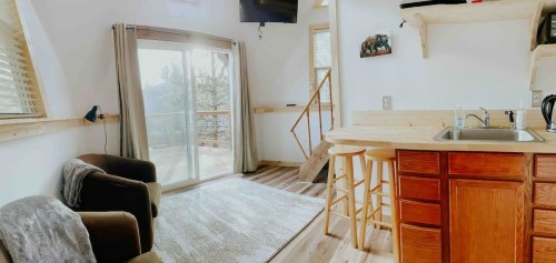 Cozy interior of a cabin with a kitchen, seating area, and large windows overlooking a deck and nature.