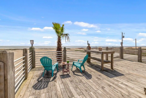 A wooden deck with two teal chairs, a palm tree, and a table overlooking a sandy beach and blue sky.